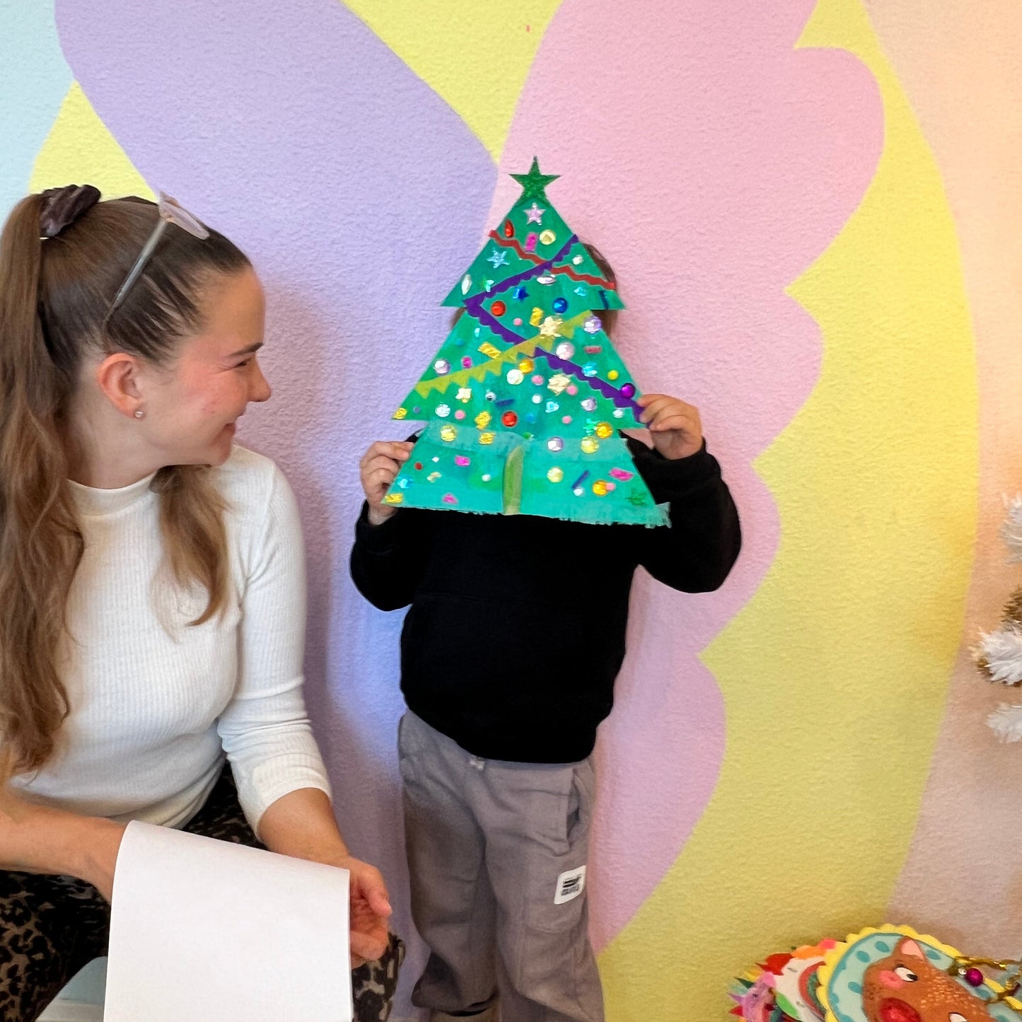 Child holding a decorated paper Christmas tree in front of a colorful butterfly cutout with a woman sitting next to them.