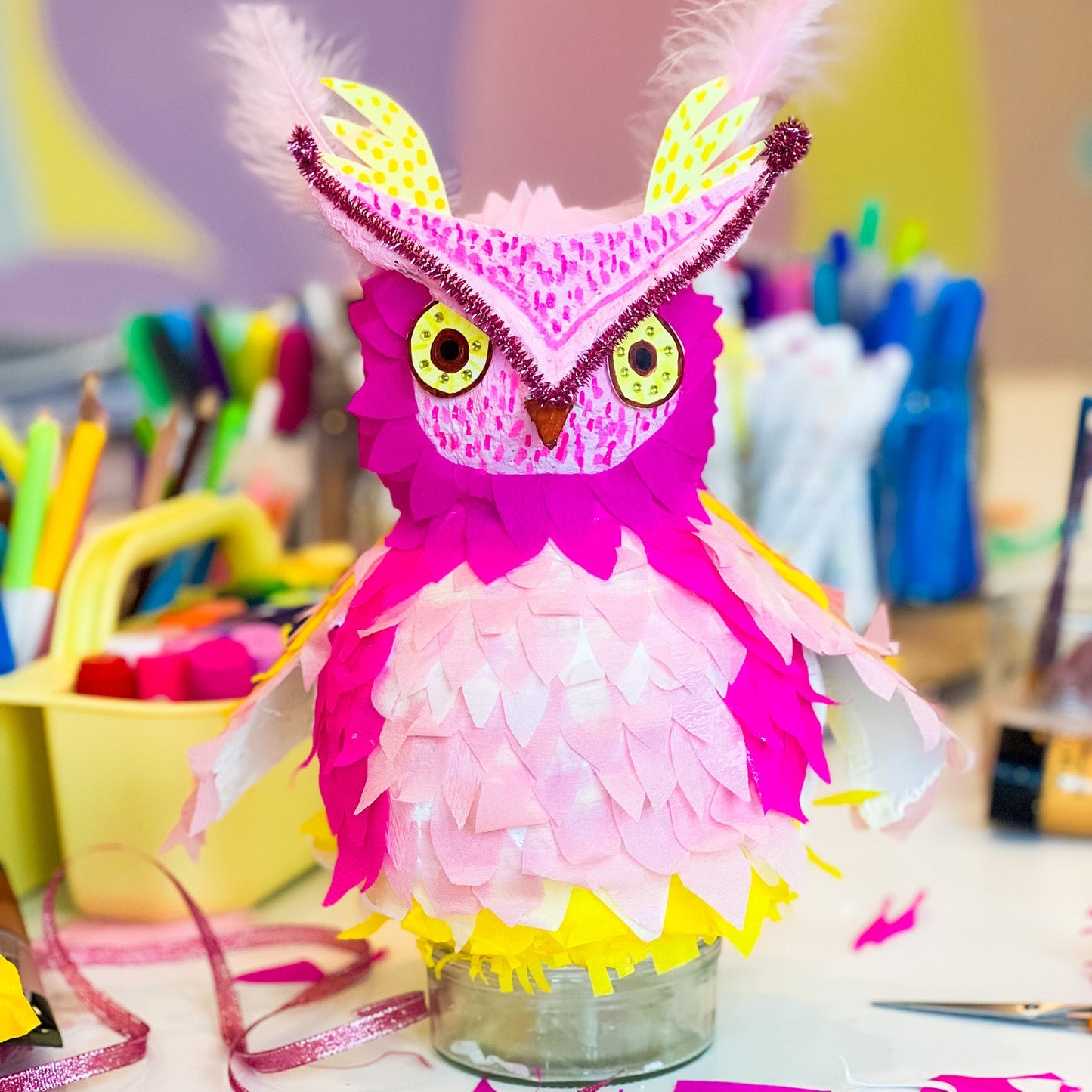 Colorful owl-shaped piñata with feathers on a table with art supplies in the background